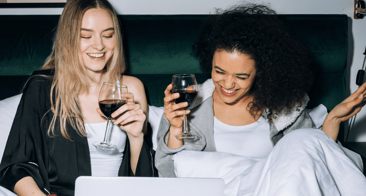 Two women drinking red wine in bed and laughing at something on a laptop