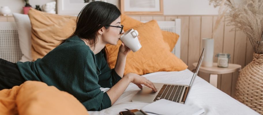 Woman lying on bed sipping coffee and working on a laptop