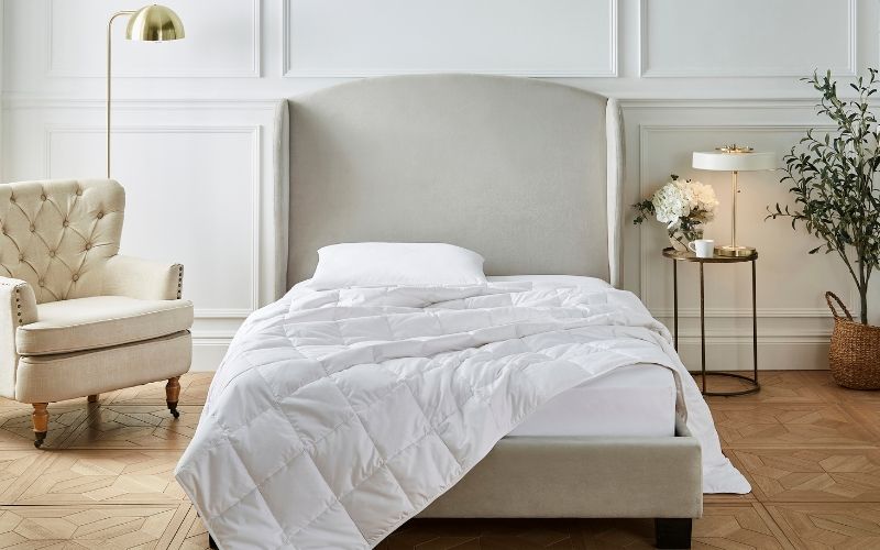 A Liddell cashmere duvet draped across a mattress, half hanging off. It's in a contemporary bedroom with a huge headboard. A lamp and a plant sits to one side. Another lamp and a chair is off to the other side. 