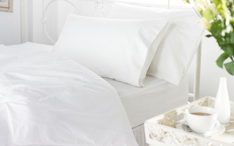 A V144 50/50 polycotton plain duvet cover on a bed, complete with pillows. A plant and cup of coffee is visible on a table next to the bed.