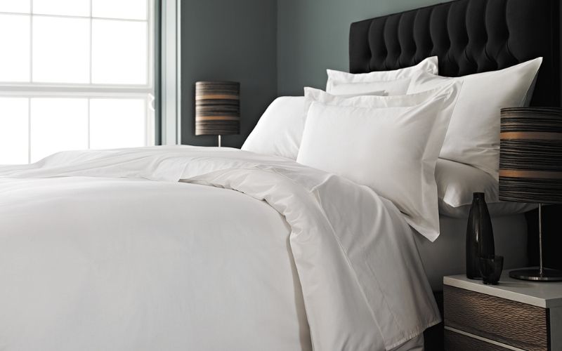 A VV300 100% cotton sateen plain duvet cover and pillowcases in a contemporary bedroom. Lamps with brown shades are featured on either side of the bed, and the headboard is black suede.